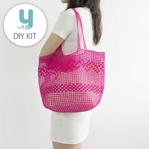 Puede incluir: Un bolso tote rosa brillante, de tejido abierto, con asas largas. El bolso lo lleva una persona que viste un vestido blanco. El bolso tiene forma rectangular con un patrón tejido. La imagen también incluye un círculo blanco con el texto "DIY KIT" y un logotipo "y" estilizado.