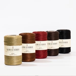 Op de afbeelding: Vijf spoelen leer draad in verschillende kleuren: beige, bruin, rood, donkerbruin en zwart. Elke spoel heeft een wit etiket met de tekst "LIM LEATHER" en "SLIM LEATHER". De draad is strak gewikkeld en lijkt van polyester te zijn.