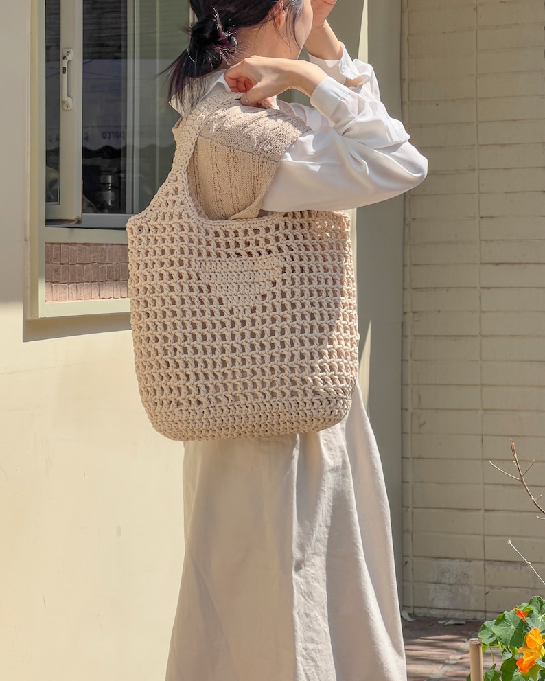Puede incluir: Una bolsa de mano de crochet beige con un bolsillo cuadrado en la parte delantera. La bolsa la lleva una persona que lleva una camisa blanca y un jersey beige.