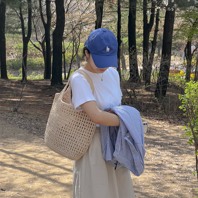 Puede incluir: Una mujer con una camiseta blanca, una gorra de b&eacute;isbol azul con un logotipo blanco y un bolso de mano de crochet beige. Lleva una camisa a rayas azules y blancas en la mano.
