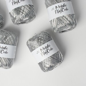 May include: Several spools of silver metallic yarn are arranged on a white surface. Each spool has a white label with the text "Metallic PlayCode" in a handwritten font. The yarn appears to be a shiny, woven material.