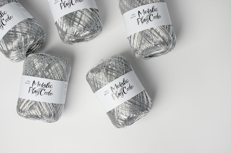 May include: Several spools of silver metallic yarn are arranged on a white surface. Each spool has a white label with the text "Metallic PlayCode" in a handwritten font. The yarn appears to be a shiny, woven material.