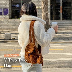 Könnte beinhalten: Ein brauner, flauschiger Rucksack mit Kordelzug. Der Rucksack wird von einer Person getragen, die eine weiße, flauschige Jacke und hellblaue Jeans trägt. Der Text "V Two Two Bag" ist im Bild sichtbar.