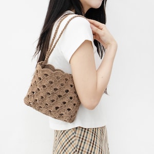 May include: A brown crocheted tote bag with a scalloped top and a woven design. The bag is being worn by a person wearing a white t-shirt and a plaid skirt.
