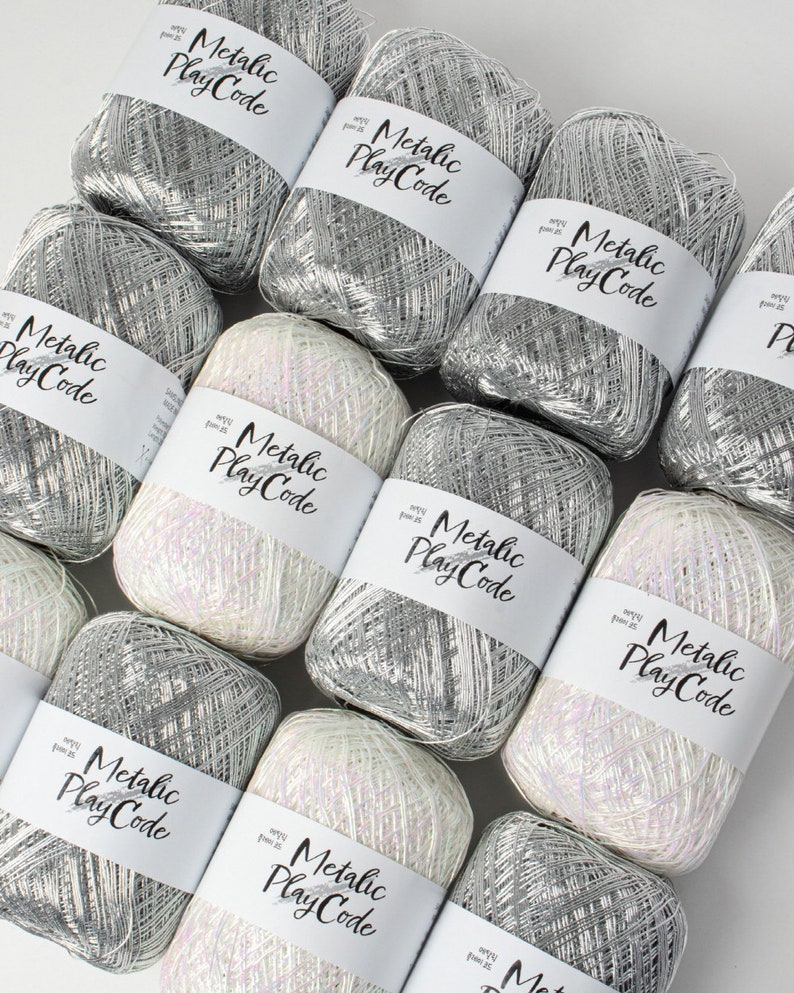 May include: An overhead shot of several skeins of metallic yarn. The yarn is silver and white, with some skeins having a pearlescent sheen. Each skein has a white label with the words "Metallic Play Code" in a handwritten font.