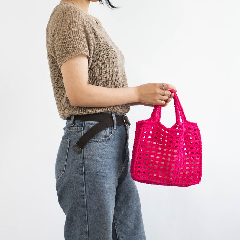 May include: A woman wearing a brown sweater and blue jeans is holding a bright pink crocheted tote bag.