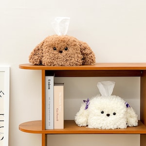 Puede incluir: Dos fundas de caja de pañuelos de peluche con forma de perro. El perro marrón tiene una textura esponjosa y el perro blanco tiene una textura esponjosa con lazos morados. Las fundas de caja de pañuelos están en un estante de madera con libros.