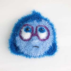 May include: A blue and white crocheted plush toy with a sad expression. The toy has purple glasses and a frown.