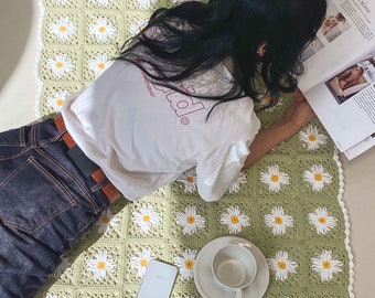 May include: A green crocheted blanket with a daisy pattern. A person is lying on the blanket reading a magazine. A white phone and a white cup are on the blanket.