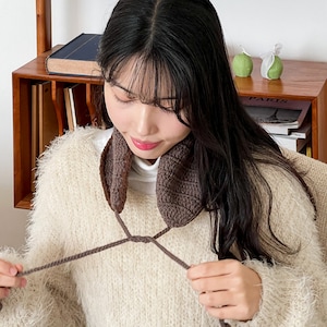 May include: A brown crocheted neck warmer with a long, thin cord that ties at the front. The neck warmer is being worn by a person with long, dark hair.