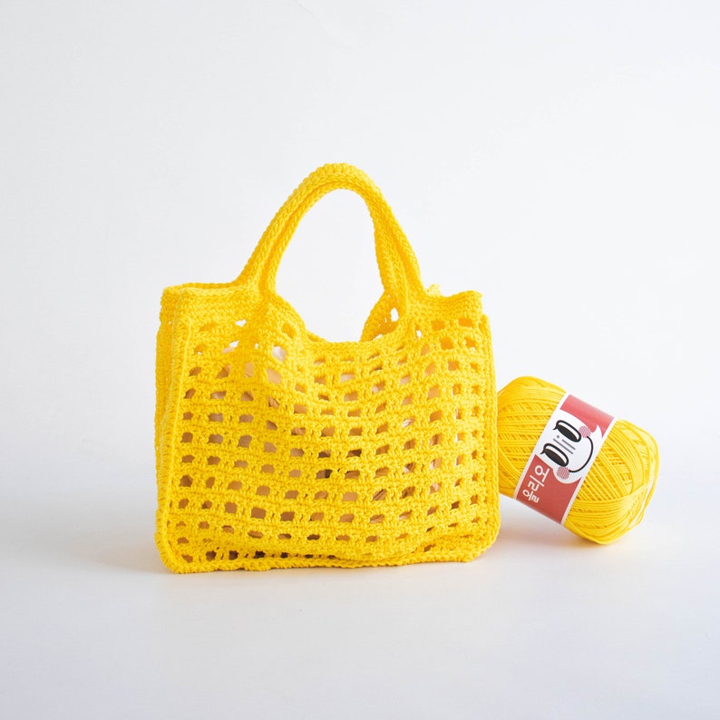 May include: A bright yellow crochet tote bag with a wide, open weave. The bag has a single handle and is sitting next to a ball of yellow yarn with a red label that says "D.I.Y.  TOTE"