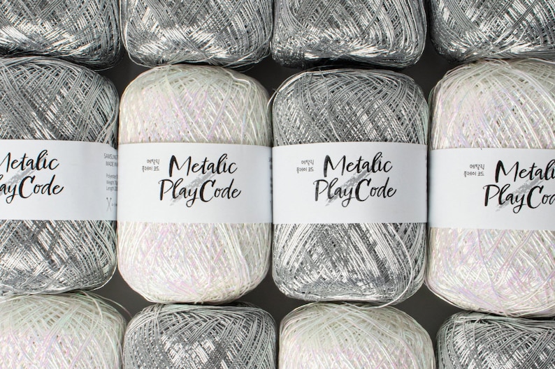 May include: Close-up of several spools of metallic yarn. The yarn is available in silver and white, with a shimmering, iridescent quality. Each spool has a white label with the text "Metallic PlayCode". The yarn is tightly wound and appears soft.