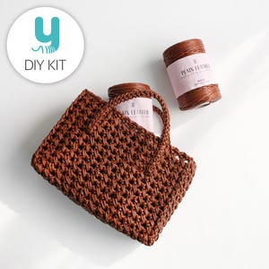 May include: A brown woven tote bag with two handles, accompanied by two spools of brown leather cord. The bag and cord are part of a DIY kit, as indicated by the "DIY KIT" text and logo in the upper left corner.