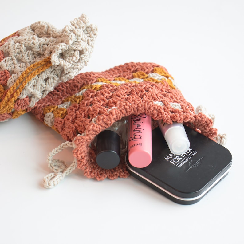 May include: A crocheted pouch with a drawstring closure, filled with makeup and beauty products. The pouch is made with a beige and brown yarn and has a textured pattern. The products include a pink lip gloss, a clear bottle with a black cap, and a black compact with the text "MAKE FOR EVER PROFESSIONAL - PARIS".
