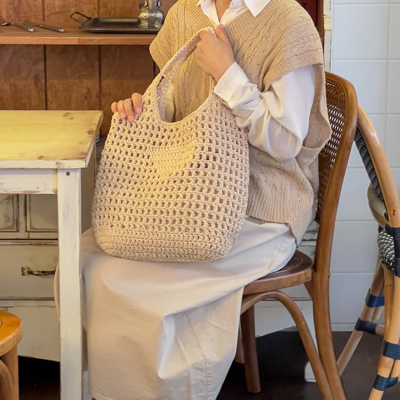 Puede incluir: Un bolso tote de crochet beige con una parte superior abierta grande y un solo asa. El bolso est&aacute; hecho de un hilo grueso y texturizado y tiene un tejido suelto y abierto.