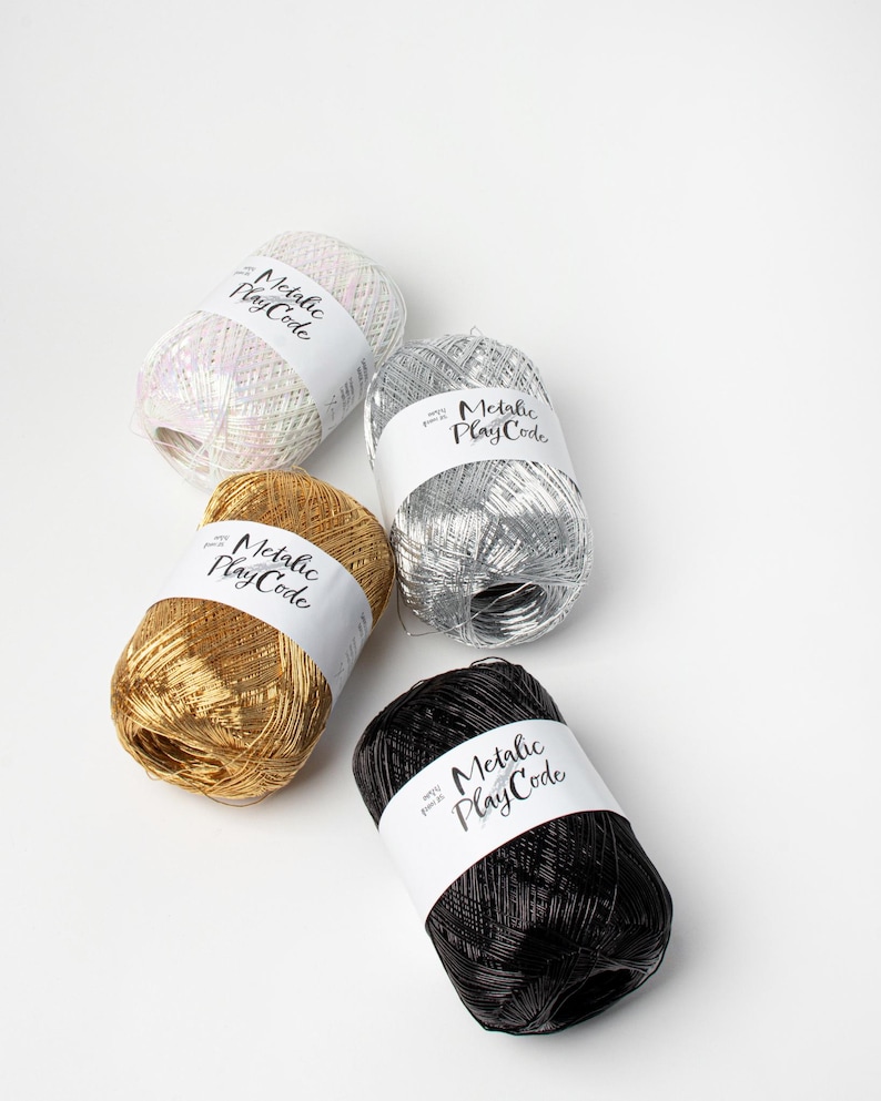May include: Four spools of metallic yarn in white, silver, gold, and black. Each spool has a white label with the text "Metallic Play Code" in a handwritten style font. The yarn is arranged on a white background.
