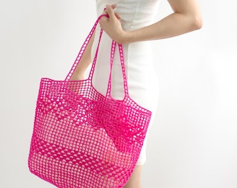 May include: A bright pink, open-weave tote bag with long handles. The bag is held by a person wearing a white dress. The bag's mesh design is visible, and it appears to be empty.