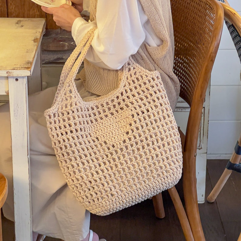Puede incluir: Un bolso tote de crochet beige con un tejido abierto grande. El bolso tiene dos asas y est&aacute; hecho de un hilo grueso y texturizado.