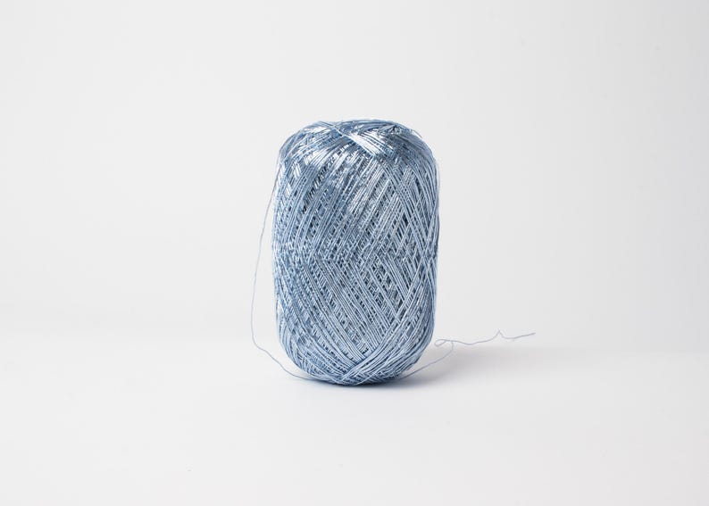 May include: A spool of light blue thread, tightly wound, sits against a white background. The thread has a metallic sheen, suggesting it may be used for decorative purposes or embroidery. A small length of thread extends from the spool.