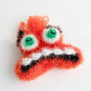 May include: A bright orange, black and white crocheted monster with green eyes. The monster has a wide, toothy grin.