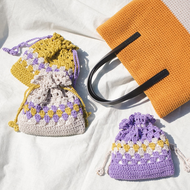 May include: Three crocheted drawstring bags in different colors. The bags are made with a square stitch pattern and feature a combination of purple, yellow, and white yarn.