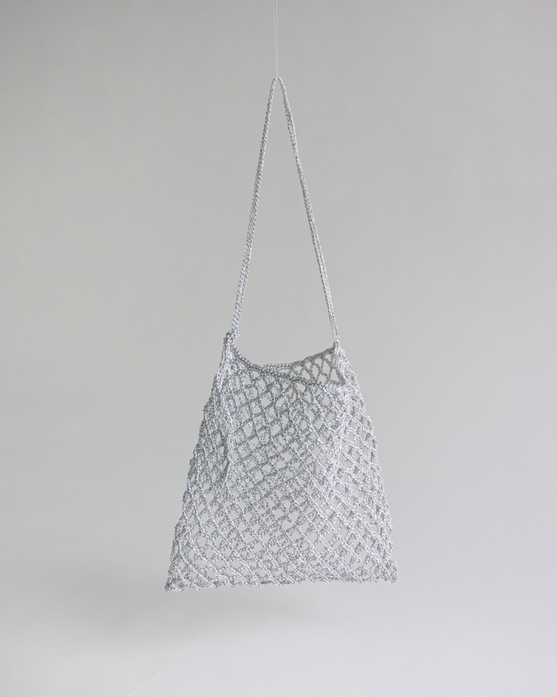 May include: A silver crocheted tote bag with a long strap, suspended against a white background. The bag features a diamond-shaped open weave design. The strap is also crocheted and appears to be attached to the top of the bag.