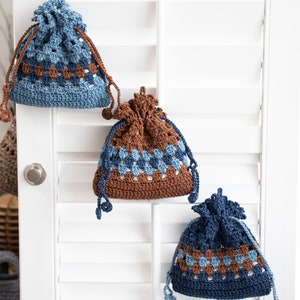 May include: Three crocheted drawstring bags with a brown and blue geometric pattern. The bags are hanging on a white shutter.