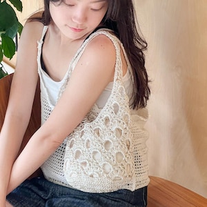 Op de afbeelding: Een wit gebreid tanktop met een groot, opengewerkt ontwerp. Het tanktop wordt gedragen door een persoon met lang donker haar.