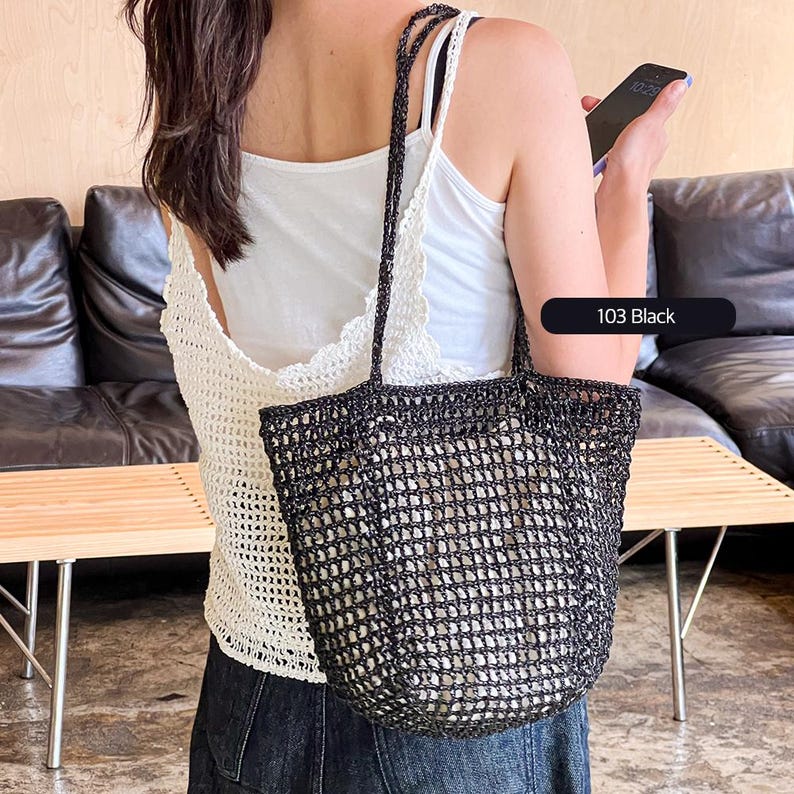 May include: A black crochet tote bag with a mesh design. The bag is being held by a person wearing a white crochet top and jeans. 103 Black