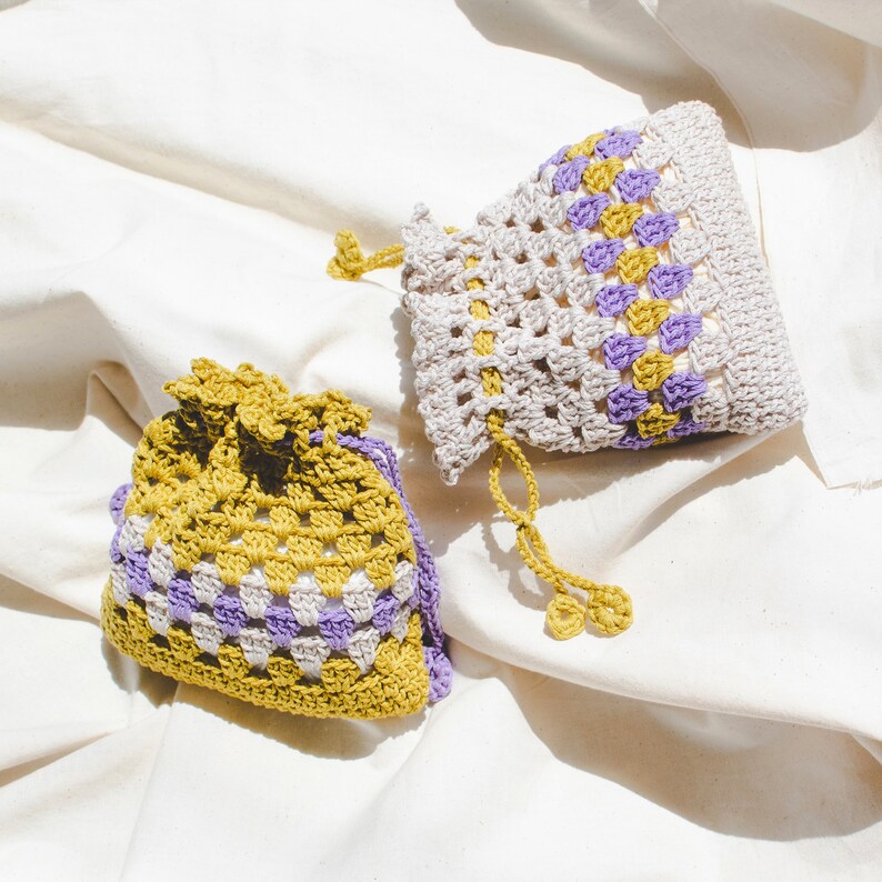 May include: Two crocheted drawstring bags with a checkered pattern in shades of beige, purple, and yellow. The bags are made with a loose, open stitch.