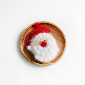 May include: A white and red knitted Santa Claus ornament with a white beard and red hat, sitting on a wooden plate.