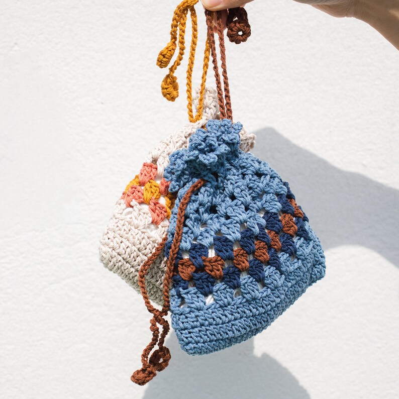 May include: Two crocheted drawstring bags, one in a light beige with orange and yellow accents, and the other in a blue with brown and white accents.