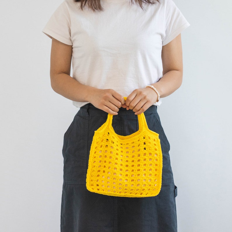 May include: A person holding a yellow crocheted tote bag. The bag has a square grid pattern.