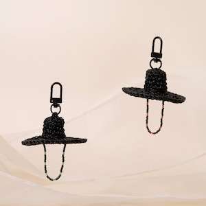 Op de afbeelding: Twee zwarte geweven hoed sleutelhangers met een kralen accent. De hoeden hebben een brede rand en een kleine kroon en zijn bevestigd aan een zwarte metalen sluiting. Het kralen accent is een U-vorm met roze, groene en zwarte kralen.