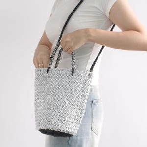 Peut inclure: Un sac fourre-tout crocheté argenté et noir avec une longue bandoulière. Le sac a un aspect texturé et tissé et une base noire. La bandoulière et les poignées sont noires et blanches. Le sac est tenu par une personne.