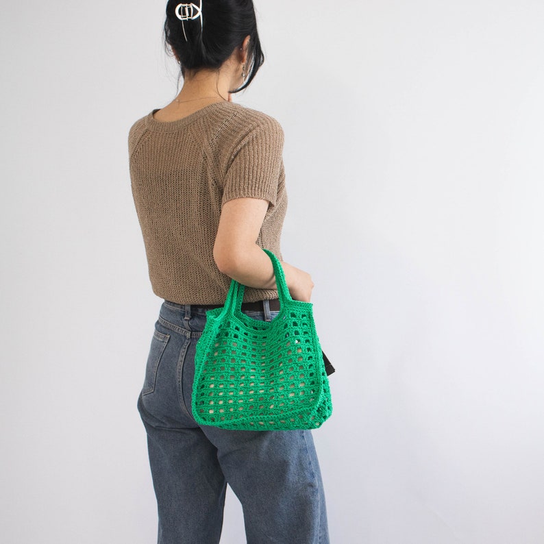 May include: A woman is holding a bright green crocheted tote bag. The bag has a square pattern and is made of thick yarn.