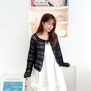 May include: A woman wearing a black crochet cardigan over a white layered dress and blue jeans. The cardigan has a loose fit and a delicate crochet pattern. The woman is standing in front of a bookshelf with books and decorative objects.