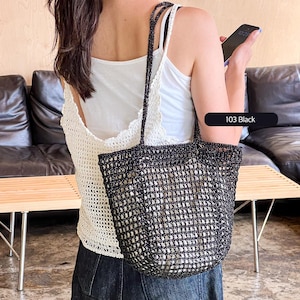 May include: Black woven tote bag with a rectangular shape and double handles. The bag has a mesh-like design, allowing visibility of the contents. The text "103 Black" is displayed on a black label. The bag is carried over the shoulder.