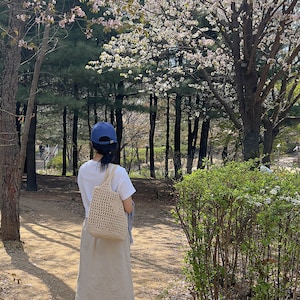 Puede incluir: Una persona con una camiseta blanca, una gorra de b&eacute;isbol azul y un bolso de mano de crochet beige camina por un parque con &aacute;rboles de cerezo en flor.