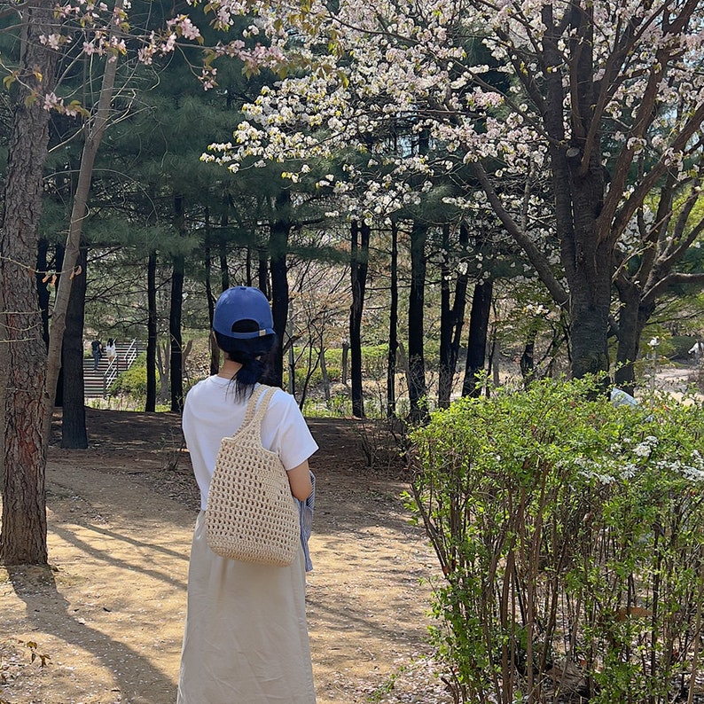 Puede incluir: Una persona con una camiseta blanca, una gorra de b&eacute;isbol azul y un bolso de mano de crochet beige camina por un parque con &aacute;rboles de cerezo en flor.