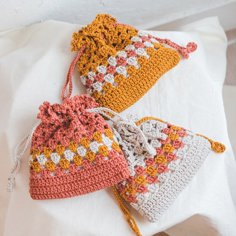 May include: Three crocheted drawstring bags in different color combinations. The bags are made with a granny square stitch and feature a mix of pink, white, and yellow yarn.