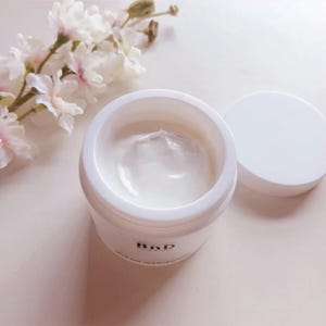 以下が含まれることがあります： 蓋が開いた白いクリームジャー。ジャーには「BnD」の文字が書かれています。クリームは滑らかでクリーミーなテクスチャーです。背景には白い花の枝があります。