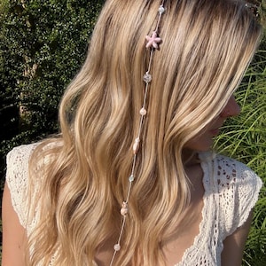 May include: A hair accessory featuring a thin white cord adorned with various beads, including a light pink starfish, clear faceted spheres, smooth white pearls, and small shell-like accents, hanging vertically against long blonde hair.