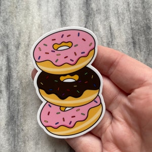 May include: A stack of three donuts, one with pink frosting and sprinkles, one with chocolate frosting and sprinkles, and one with pink frosting and sprinkles.