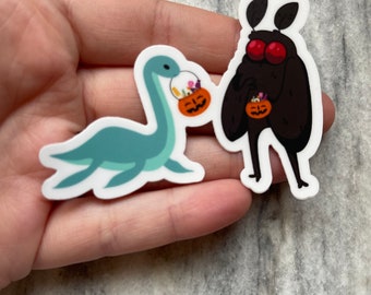Könnte beinhalten: Zwei Halloween-Sticker mit einem blauen und grünen Plesiosaurus und einem schwarzen Nachtfalter, die beide orangefarbene Kürbis-Eimer mit Süßigkeiten halten.