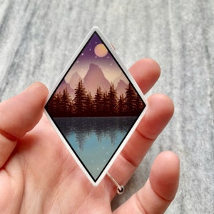 Op de afbeelding: Een sticker in de vorm van een ruit met een berglandschap. De bergen zijn paars en bruin met een blauw meer dat de lucht reflecteert. Een gele maan en sterren zijn in de lucht.