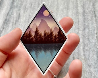 Puede incluir: Una pegatina en forma de diamante con una escena de montaña. Las montañas son moradas y marrones con un lago azul que refleja el cielo. Una luna amarilla y estrellas están en el cielo.
