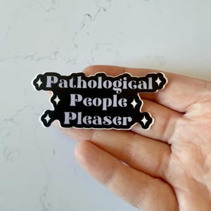 May include: A black and white sticker with the text "Pathological People Pleaser." The sticker has a unique shape with diamond accents. The sticker is held in a hand.