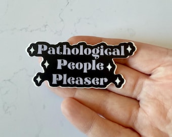 May include: A black and white sticker with the text "Pathological People Pleaser." The sticker has a unique shape with diamond accents. The sticker is held in a hand.