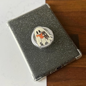 Può includere: Una custodia per tablet trasparente e glitterata con un fantasma dei cartoni animati che legge un libro con un gatto. Il fantasma è bianco con occhi neri e tiene in mano un libro arancione. Il gatto è marrone e nero.